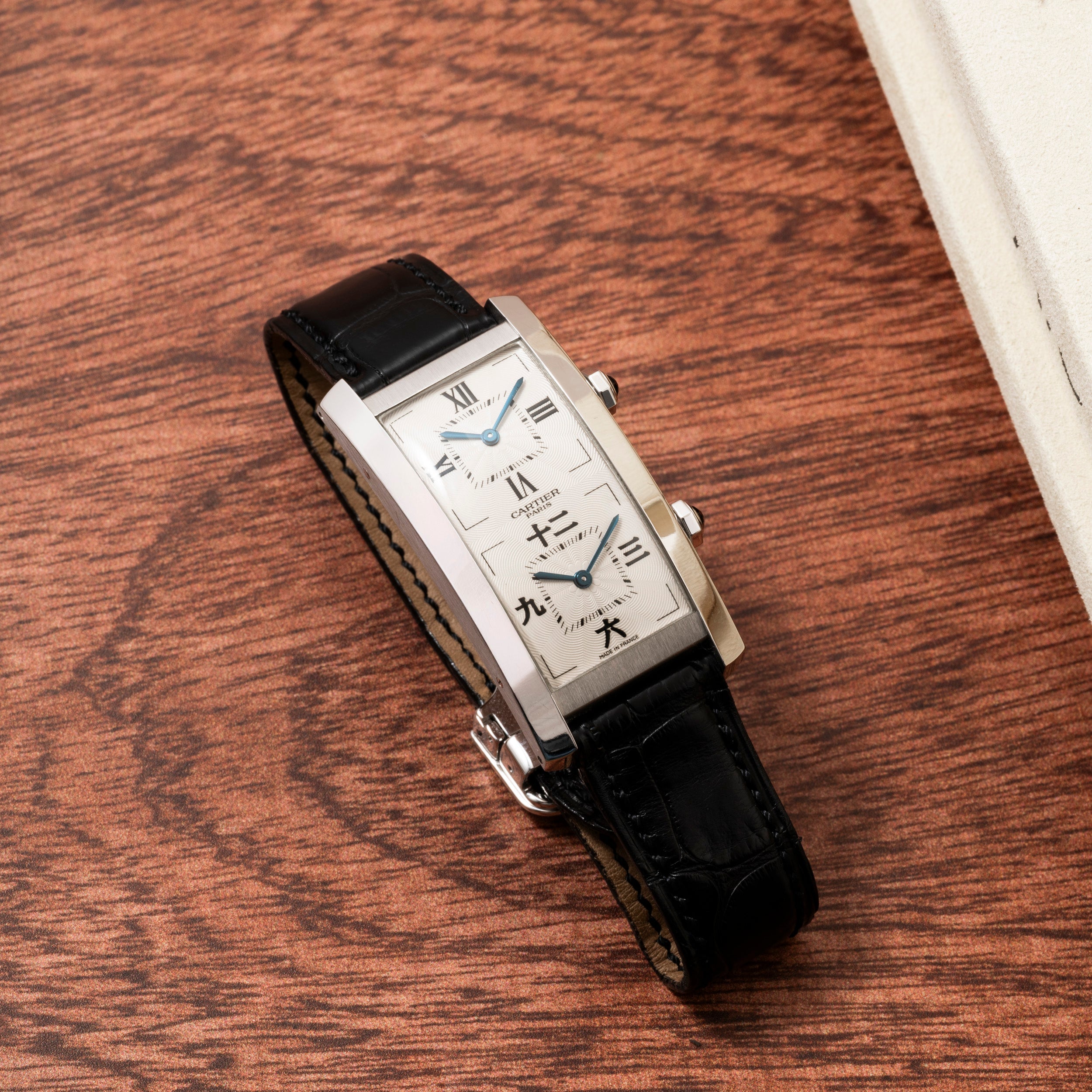 A Cartier Tank Cintrée CPCP Dual-Time rectangular silver wristwatch with a black leather strap rests on a wood surface.