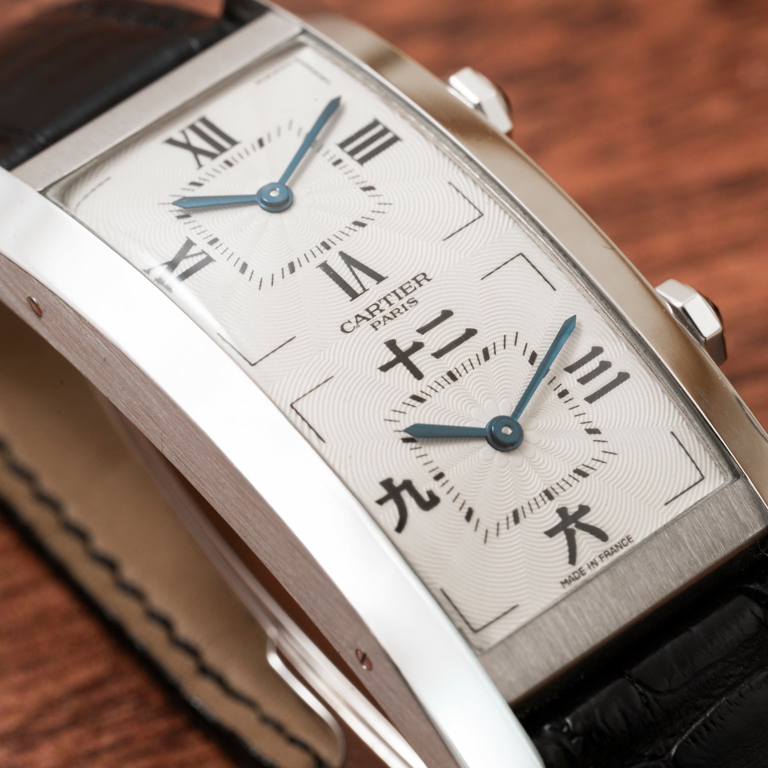 Close-up of a Cartier Tank Cintrée CPCP Dual-Time watch featuring Roman and Chinese numerals, a silver case, and a black leather strap on a wooden surface.