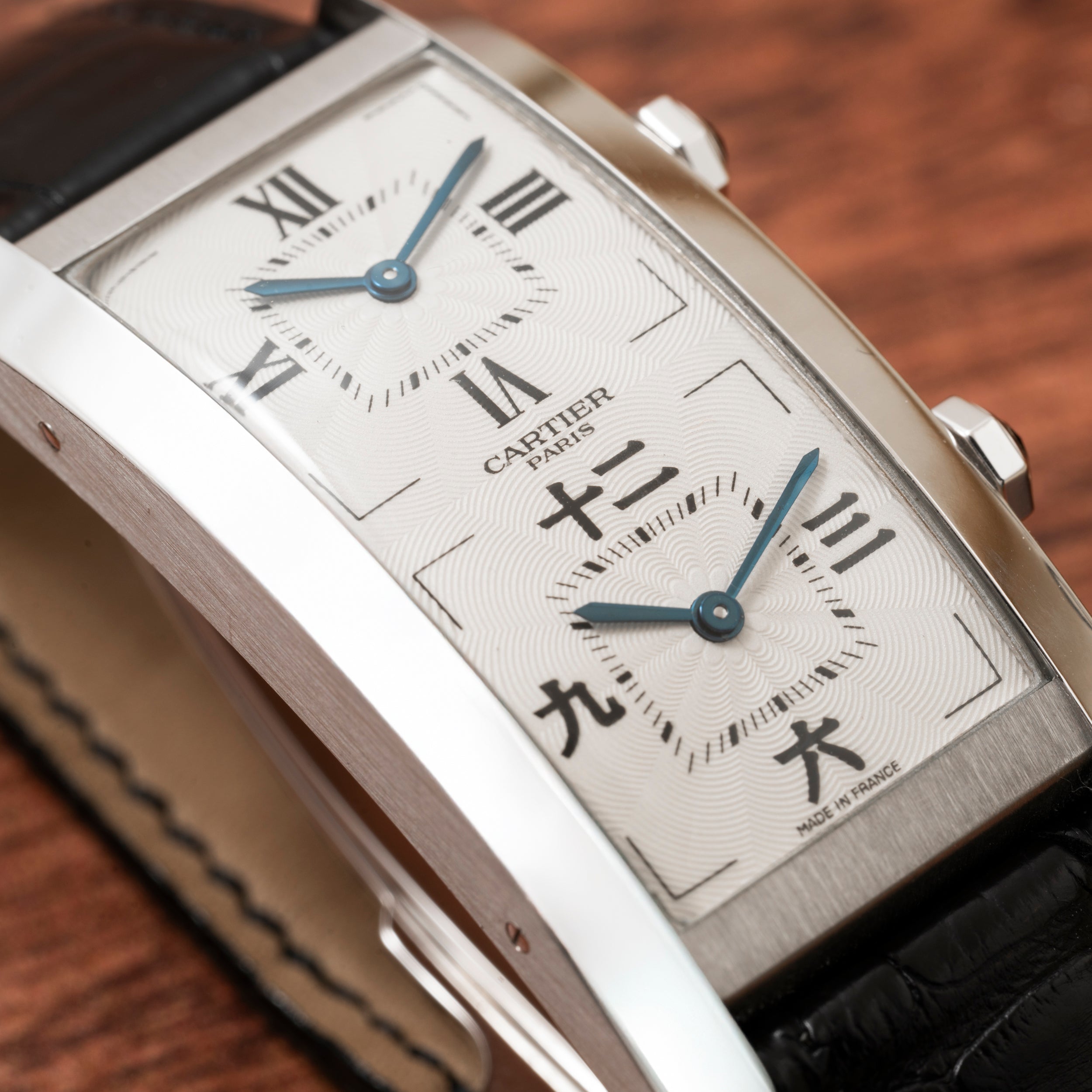Close-up of a Cartier Tank Cintrée CPCP Dual-Time watch featuring Roman and Chinese numerals, a silver case, and a black leather strap on a wooden surface.