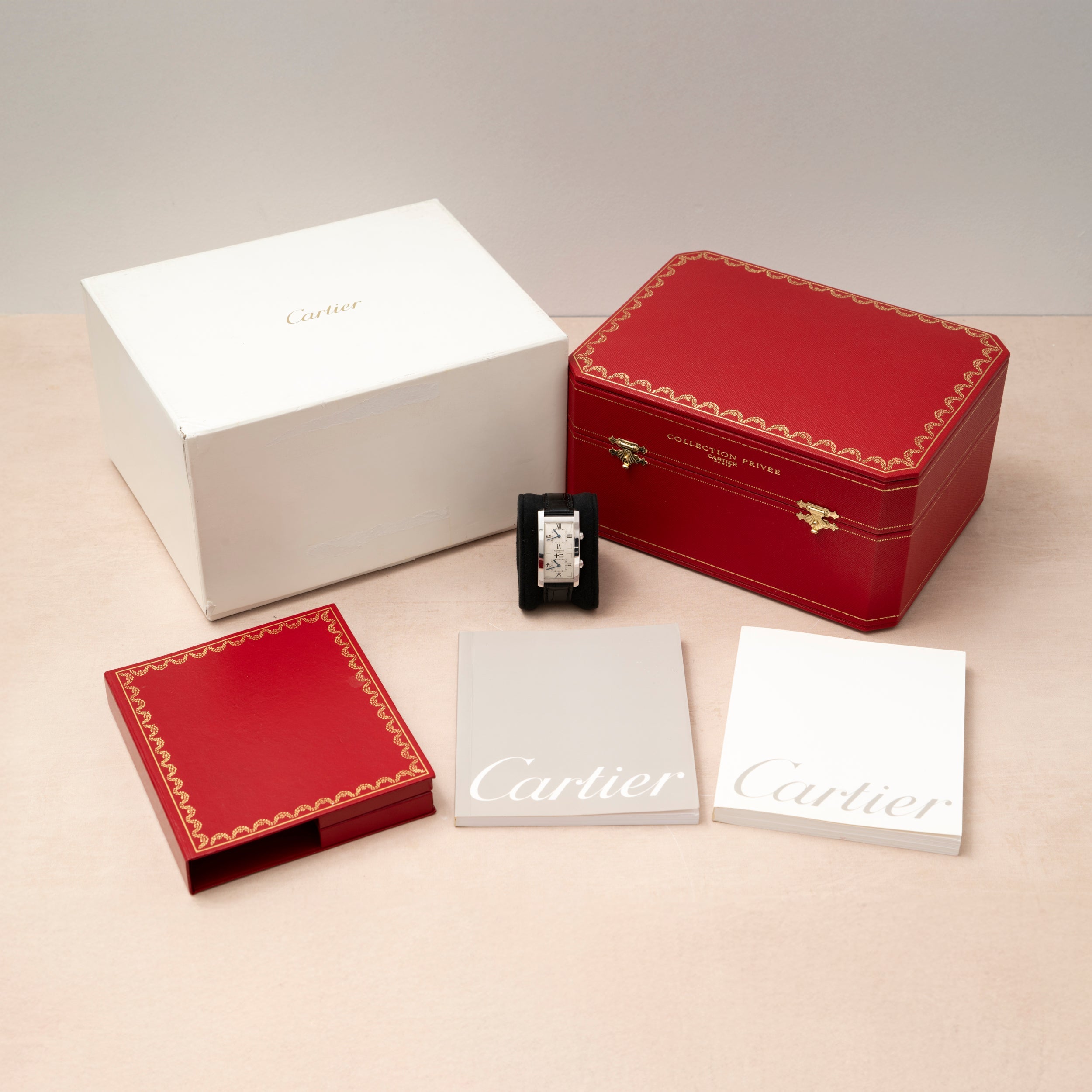 Cartier Tank Cintrée CPCP Dual-Time watch with red case, white box, and two booklets arranged neatly on a beige surface.