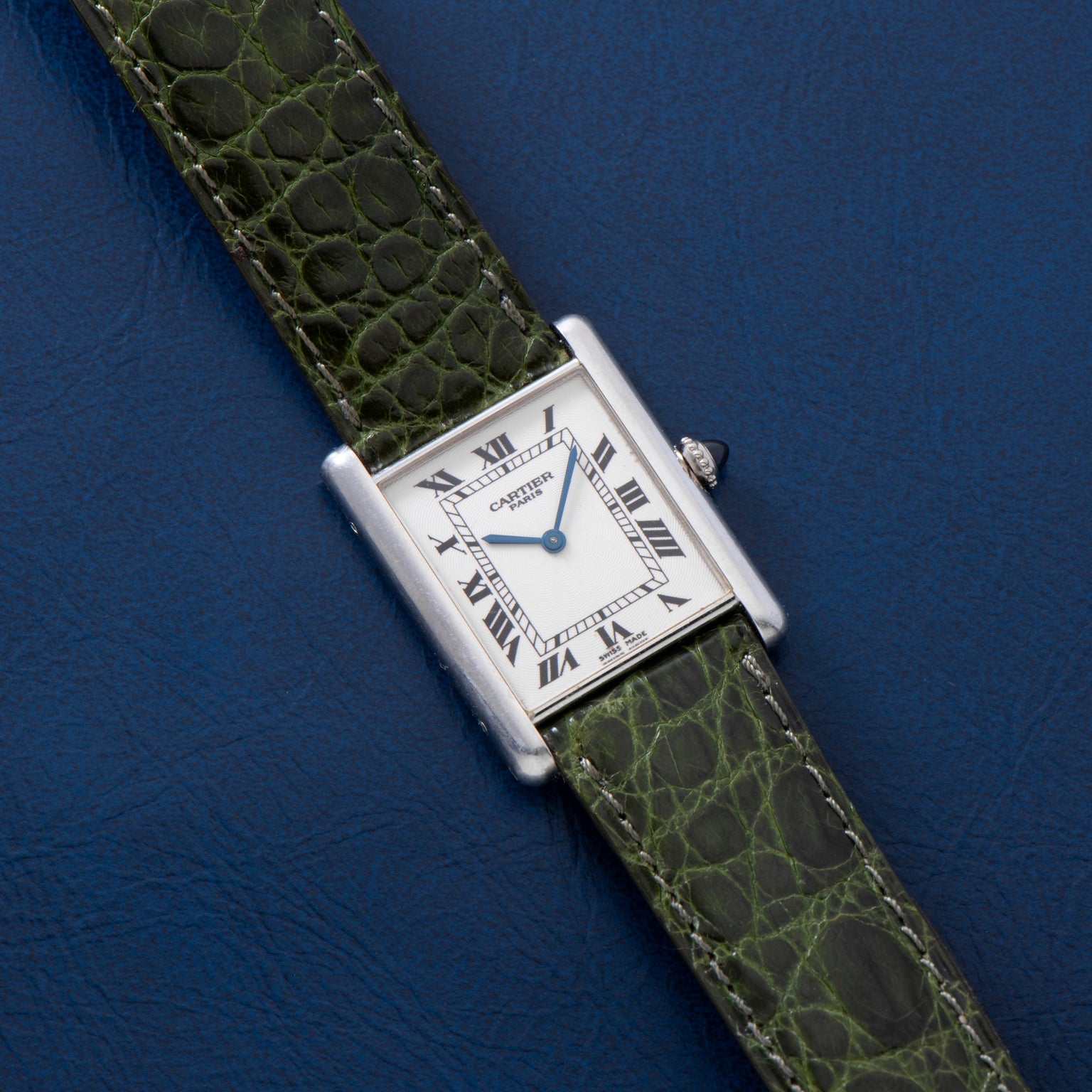 A Cartier Tank Louis CPCP watch with a green leather strap rests on a blue textured surface.