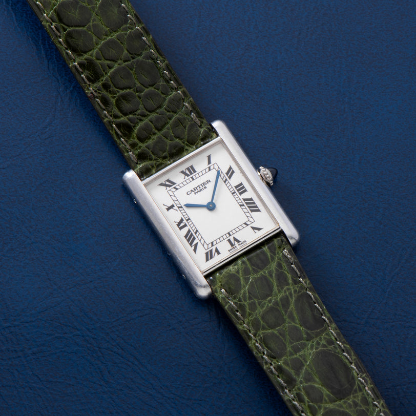 A Cartier Tank Louis CPCP watch with a green leather strap rests on a blue textured surface.
