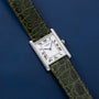 A Cartier Tank Louis CPCP watch with a green leather strap rests on a blue textured surface.