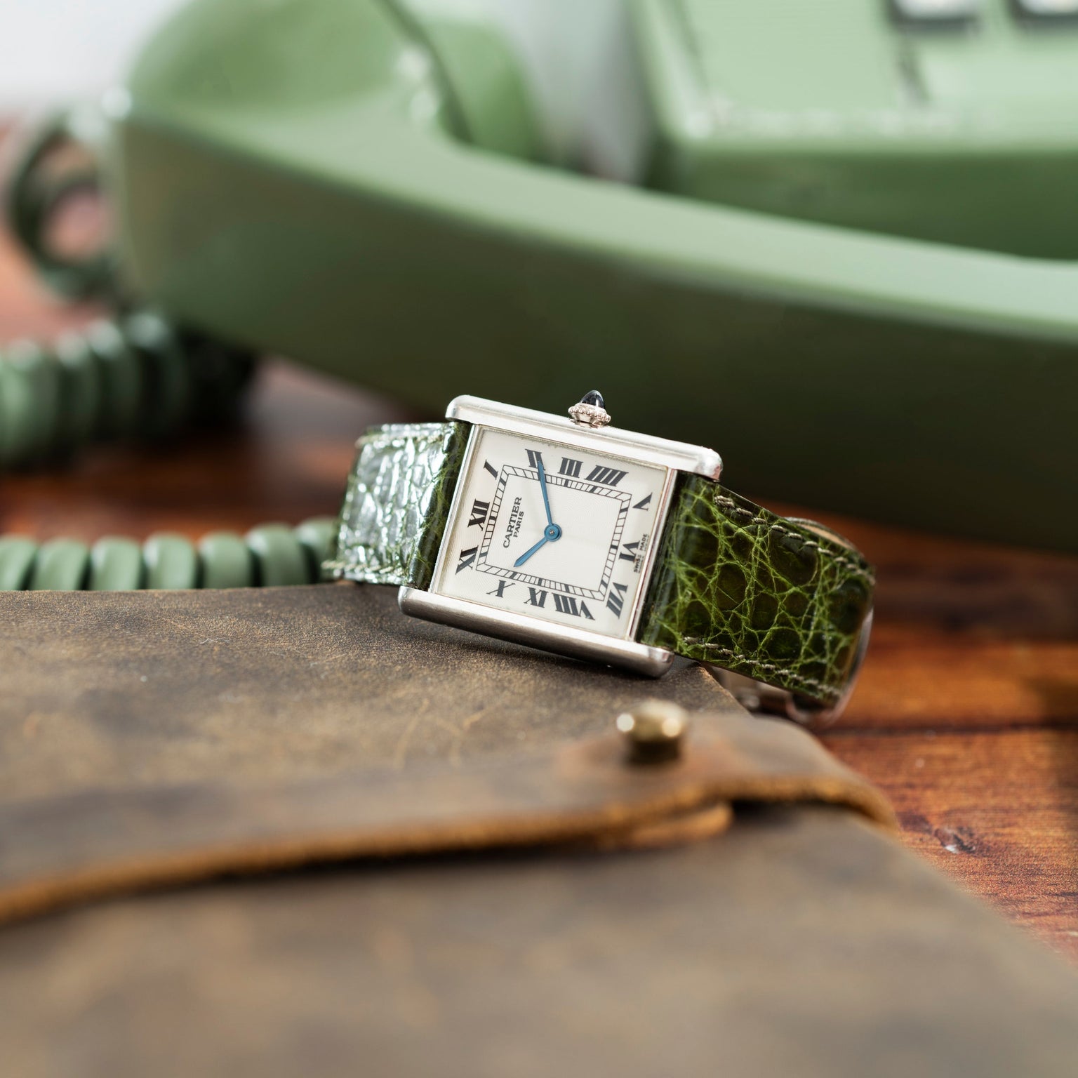 A Cartier Tank Louis CPCP rectangular wristwatch with a green leather strap sits beside a vintage green rotary phone and a brown pouch.