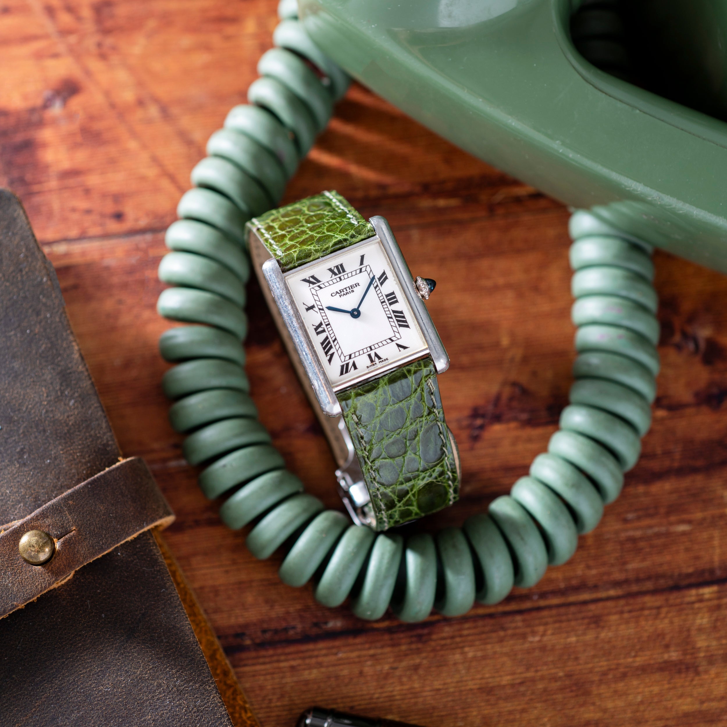 A Cartier Tank Louis CPCP wristwatch with a green leather strap rests on a coiled green phone cord atop a wooden surface.