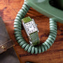 A Cartier Tank Louis CPCP wristwatch with a green leather strap rests on a coiled green phone cord atop a wooden surface.