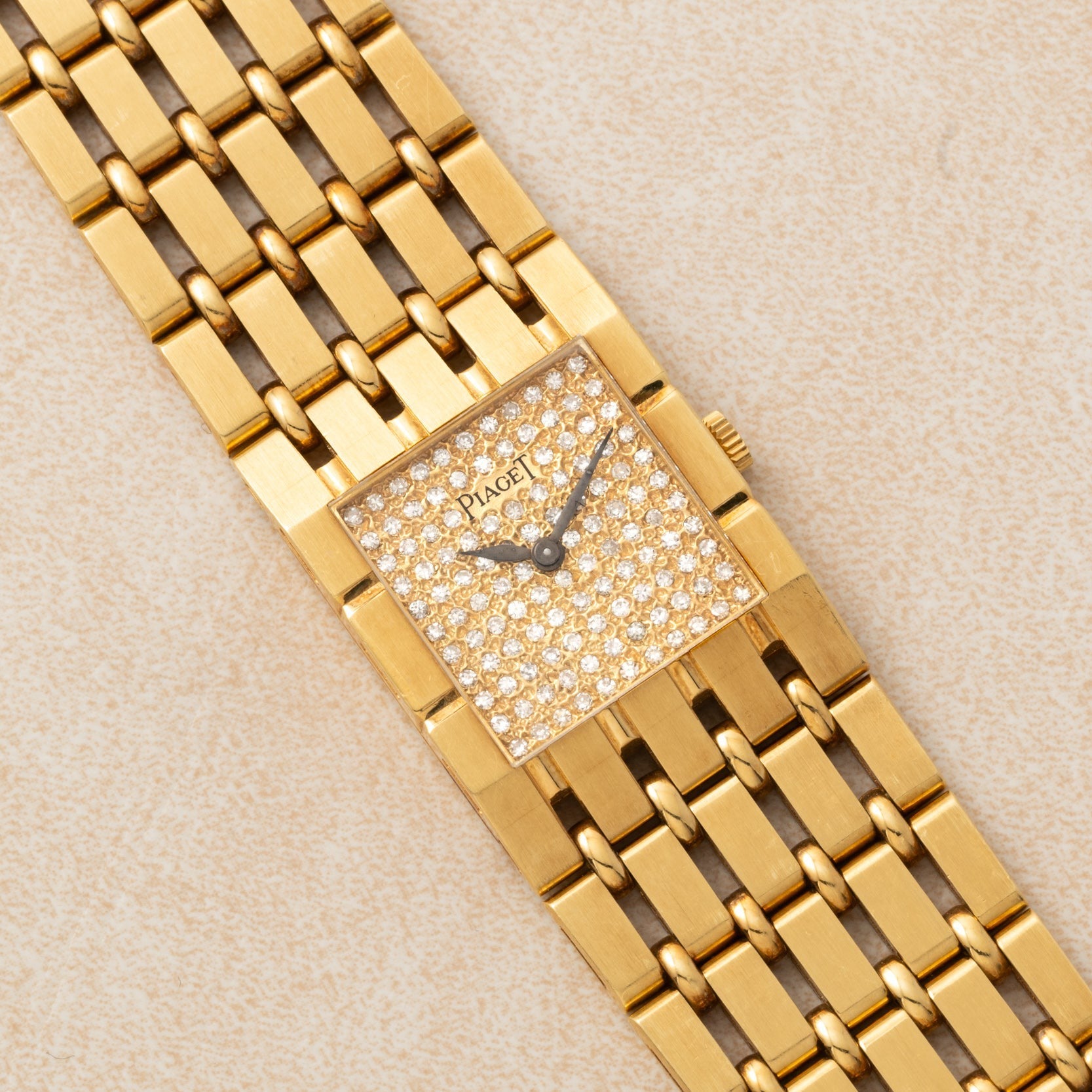 Piaget Dancer gold wristwatch featuring a rectangular, diamond-encrusted face and a linked gold bracelet, displayed on a beige surface.