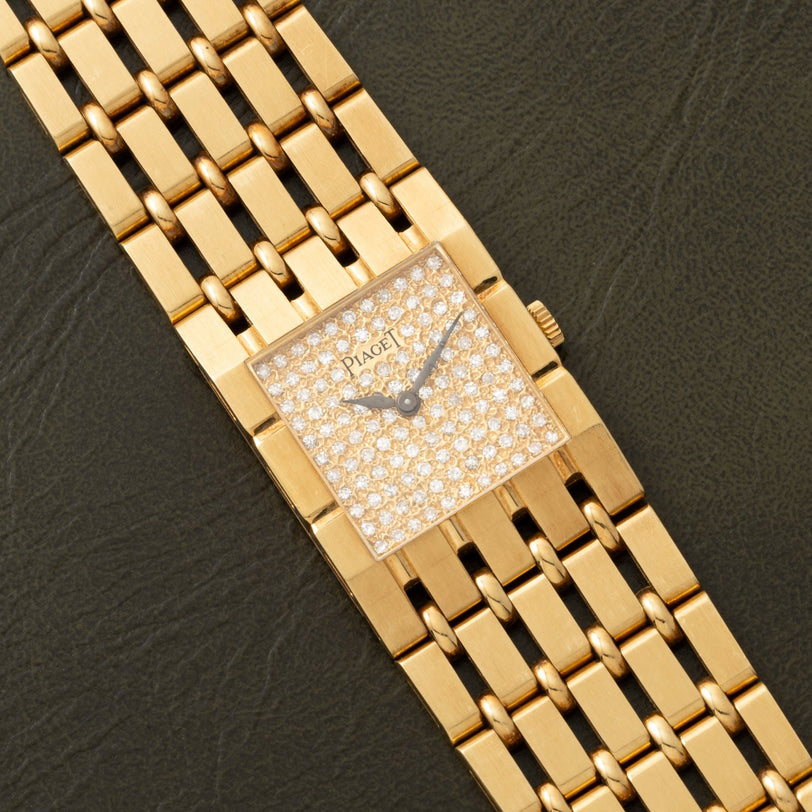 Piaget Dancer gold wristwatch with a diamond-studded square face, displayed on a dark textured surface.