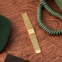 A Piaget Dancer gold wristwatch with a linked band rests on wood beside green items and a coiled phone cord.