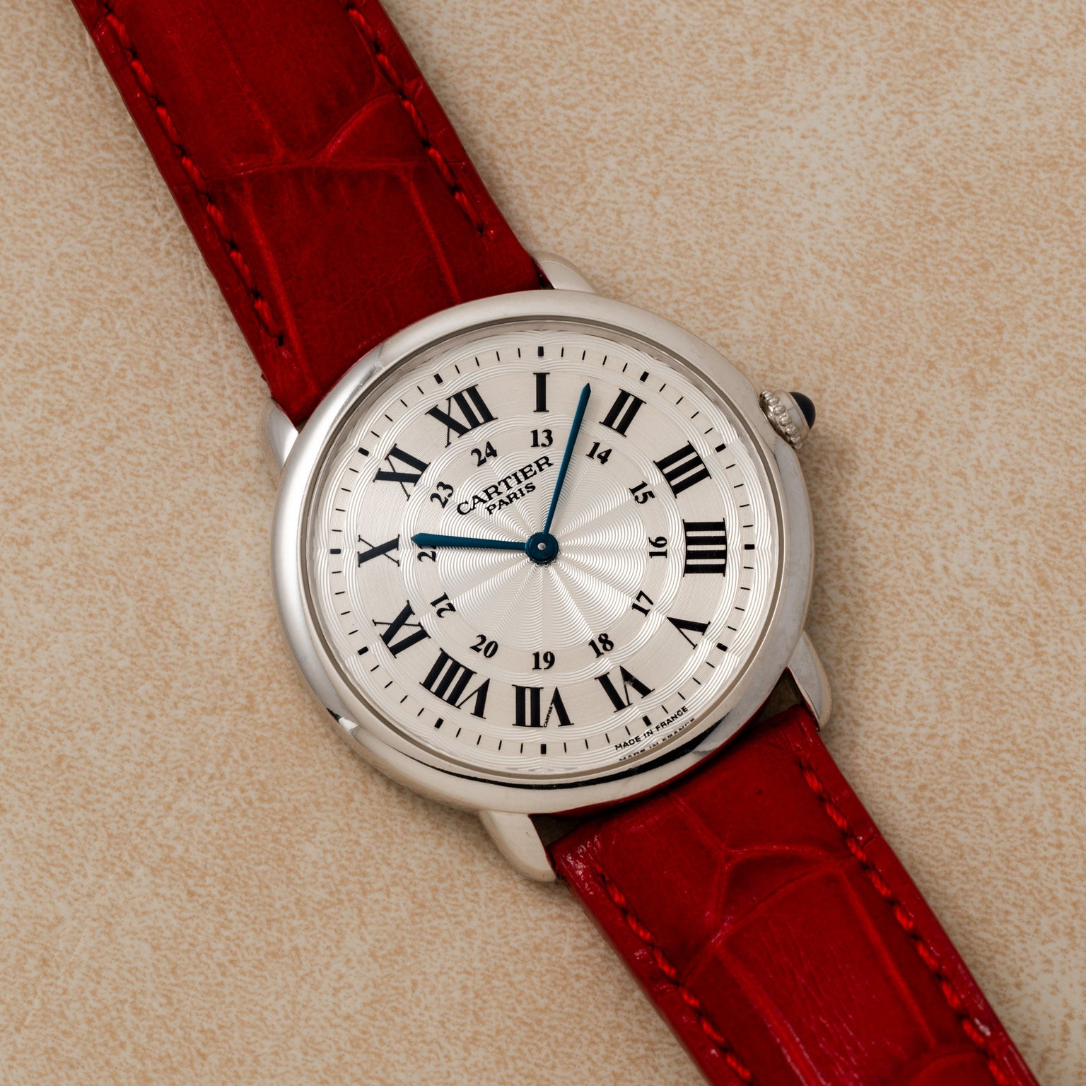 A Cartier Ronde Louis CPCP wristwatch with a red leather strap and Roman numerals rests on a beige textured surface.