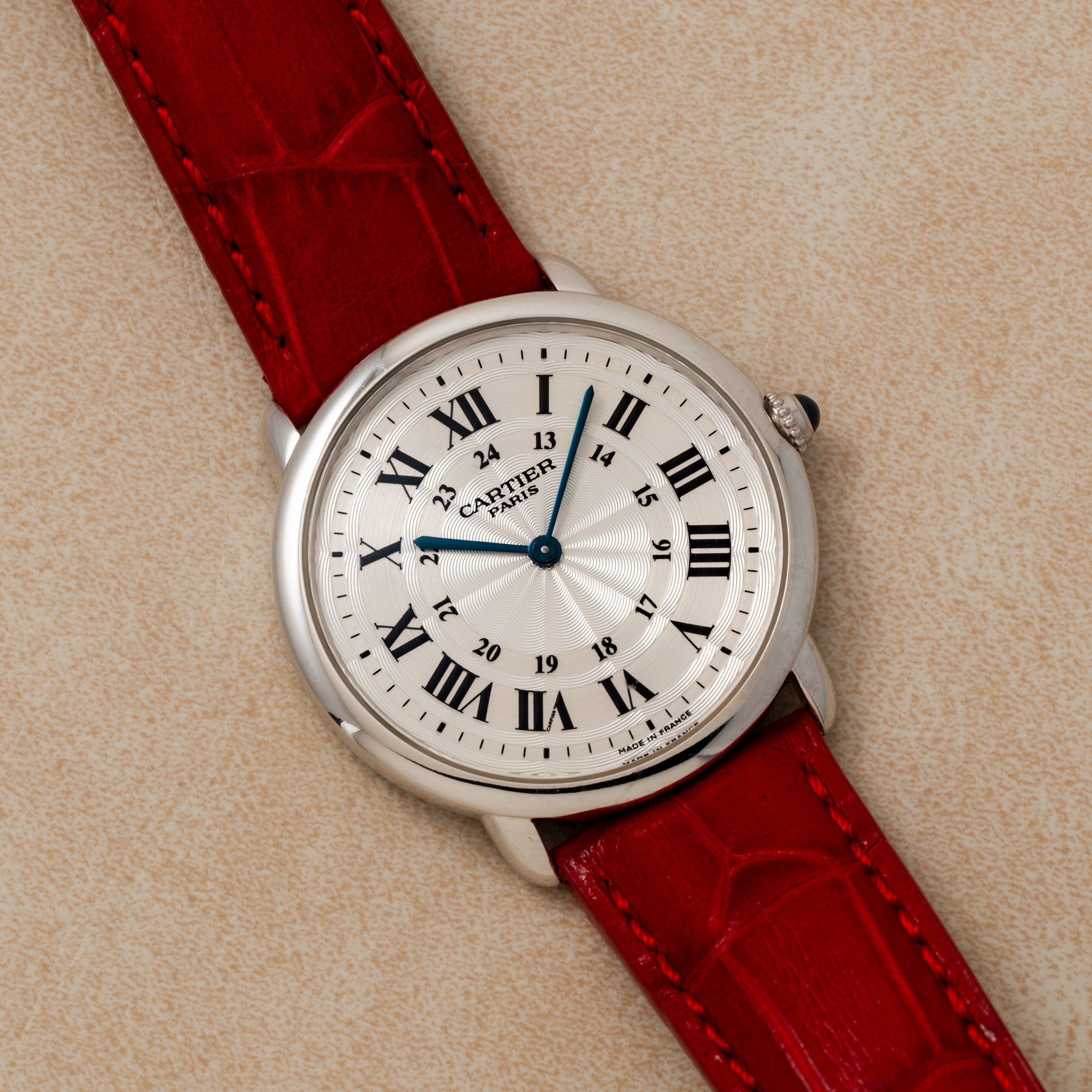 A Cartier Ronde Louis CPCP wristwatch with a red leather strap and Roman numerals rests on a beige textured surface.