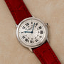 A Cartier Ronde Louis CPCP wristwatch with a red leather strap and Roman numerals rests on a beige textured surface.