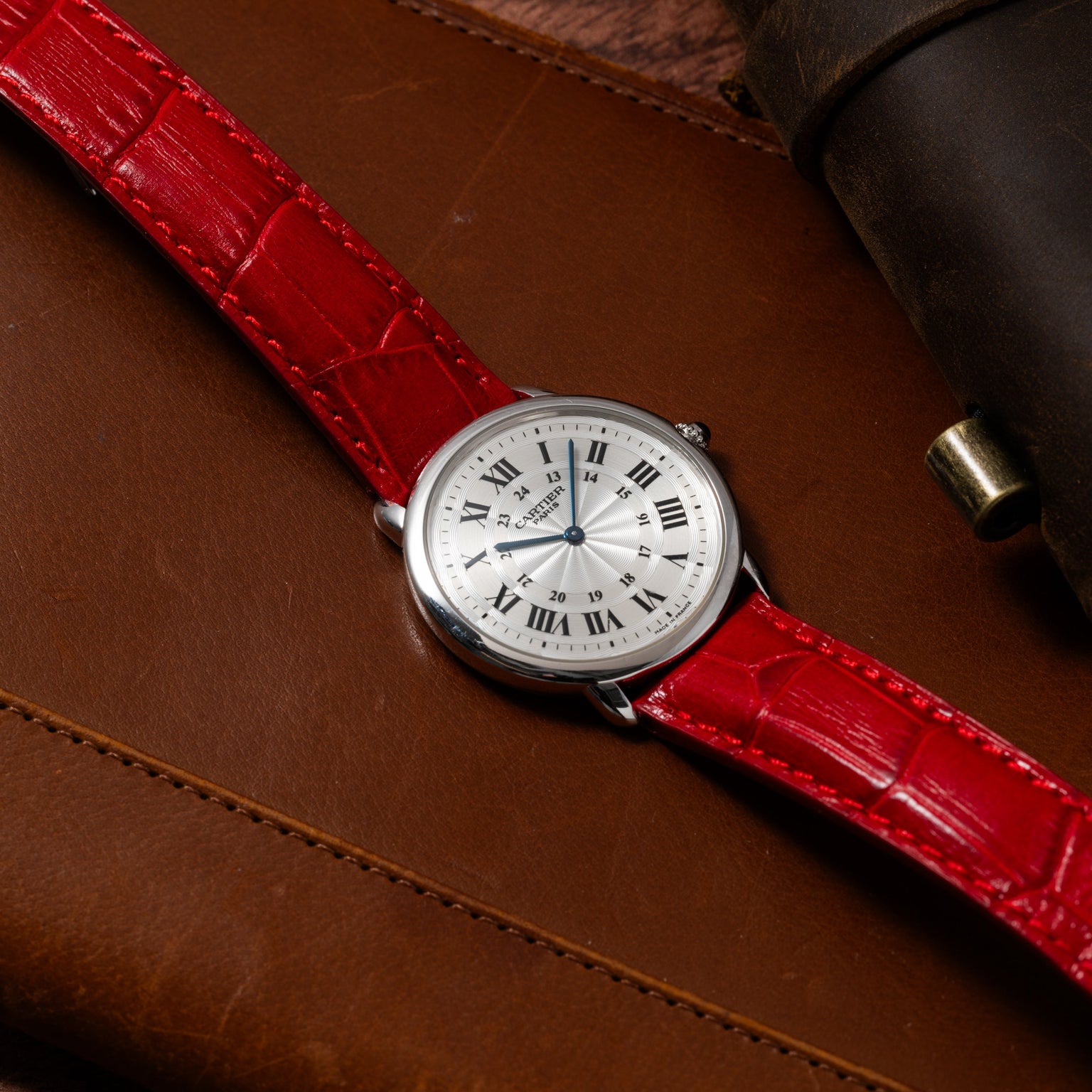 Cartier Ronde Louis CPCP watch with Roman numerals and a red leather strap displayed on a brown leather surface.