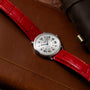 Cartier Ronde Louis CPCP watch with Roman numerals and a red leather strap displayed on a brown leather surface.