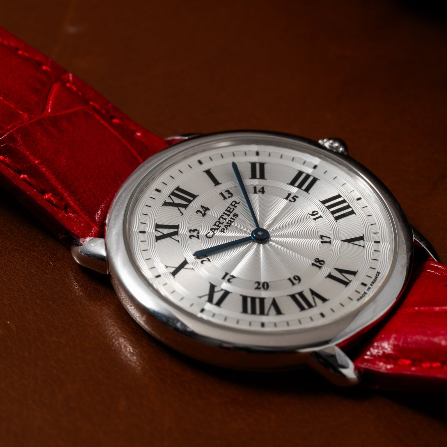 A Cartier Ronde Louis CPCP watch with a silver case and red leather strap rests on a brown surface.