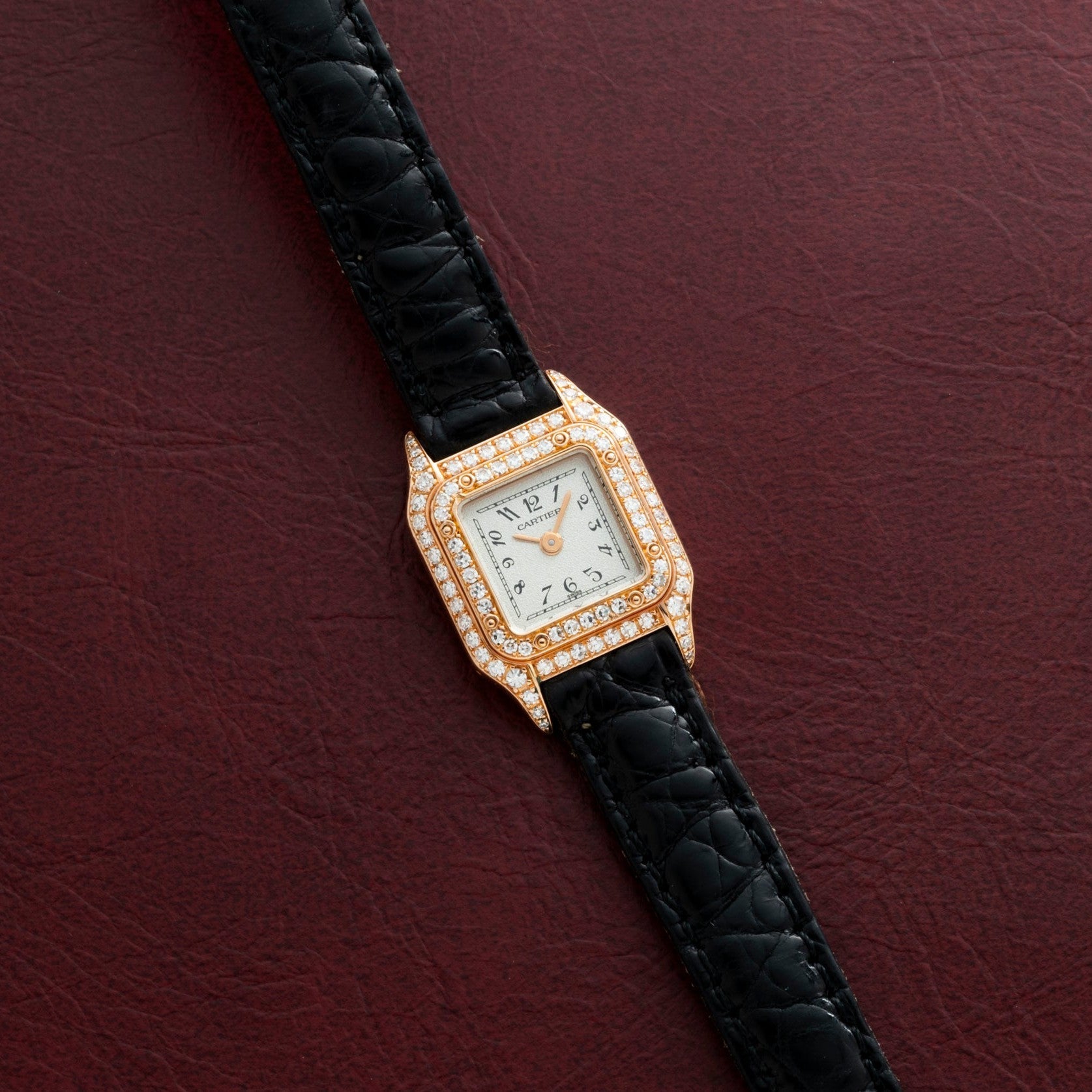 The Cartier Panthère gold watch features diamonds and a black leather strap, displayed on a textured dark red background.