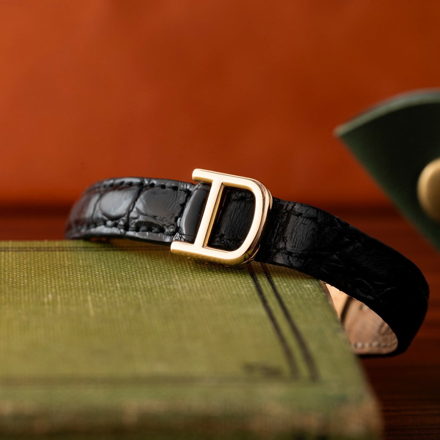 The Cartier Panthère black leather belt with a gold D buckle is displayed on a green textured book.