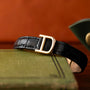 The Cartier Panthère black leather belt with a gold D buckle is displayed on a green textured book.