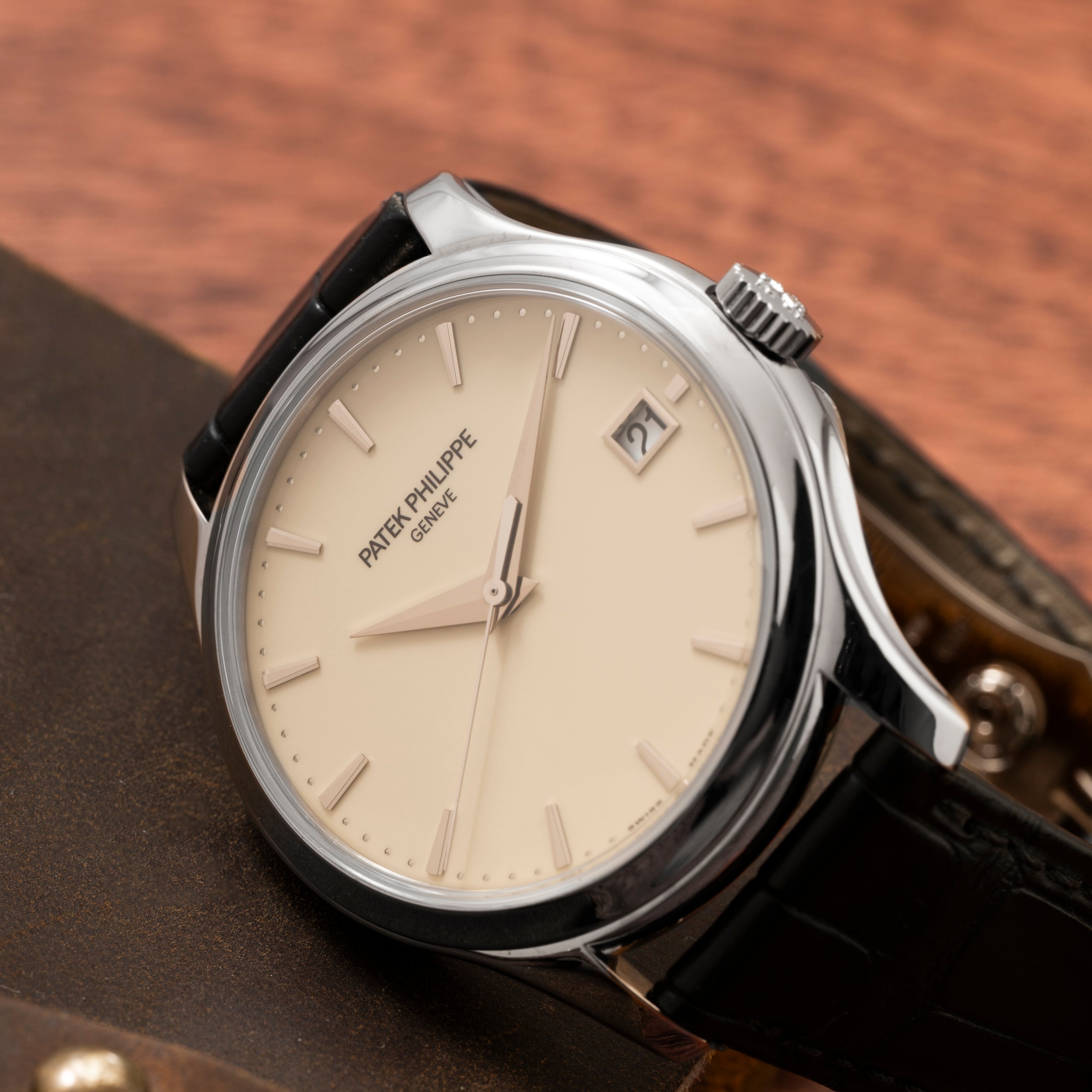 A Patek Philippe Calatrava wristwatch with a cream dial, date display, and black leather strap rests on a brown surface.
