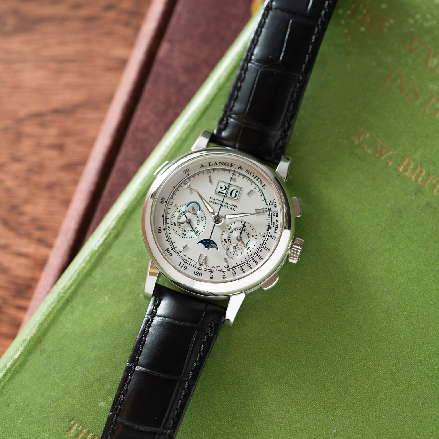 The Lange & Sohne Datograph Perpetual Calendar, a luxury wristwatch with a black leather strap, rests on a green book with gold lettering.