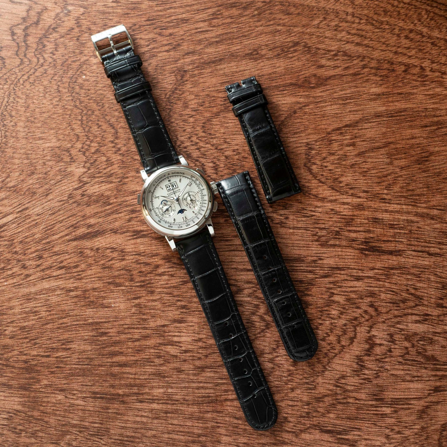 The Lange & Sohne Datograph Perpetual Calendar wristwatch with a black leather strap and two extra straps is displayed on a wooden surface.