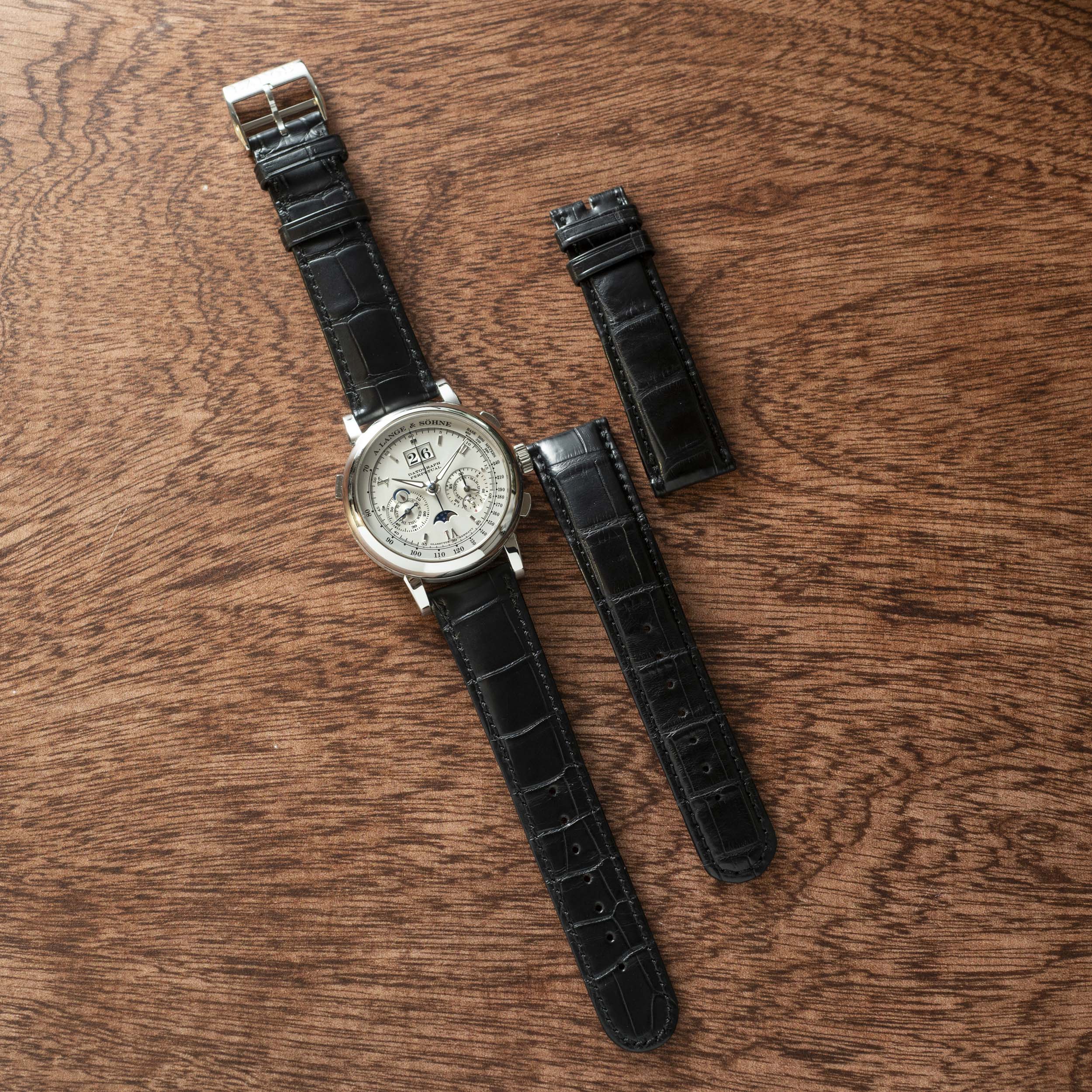 The Lange & Sohne Datograph Perpetual Calendar wristwatch with a black leather strap and two extra straps is displayed on a wooden surface.