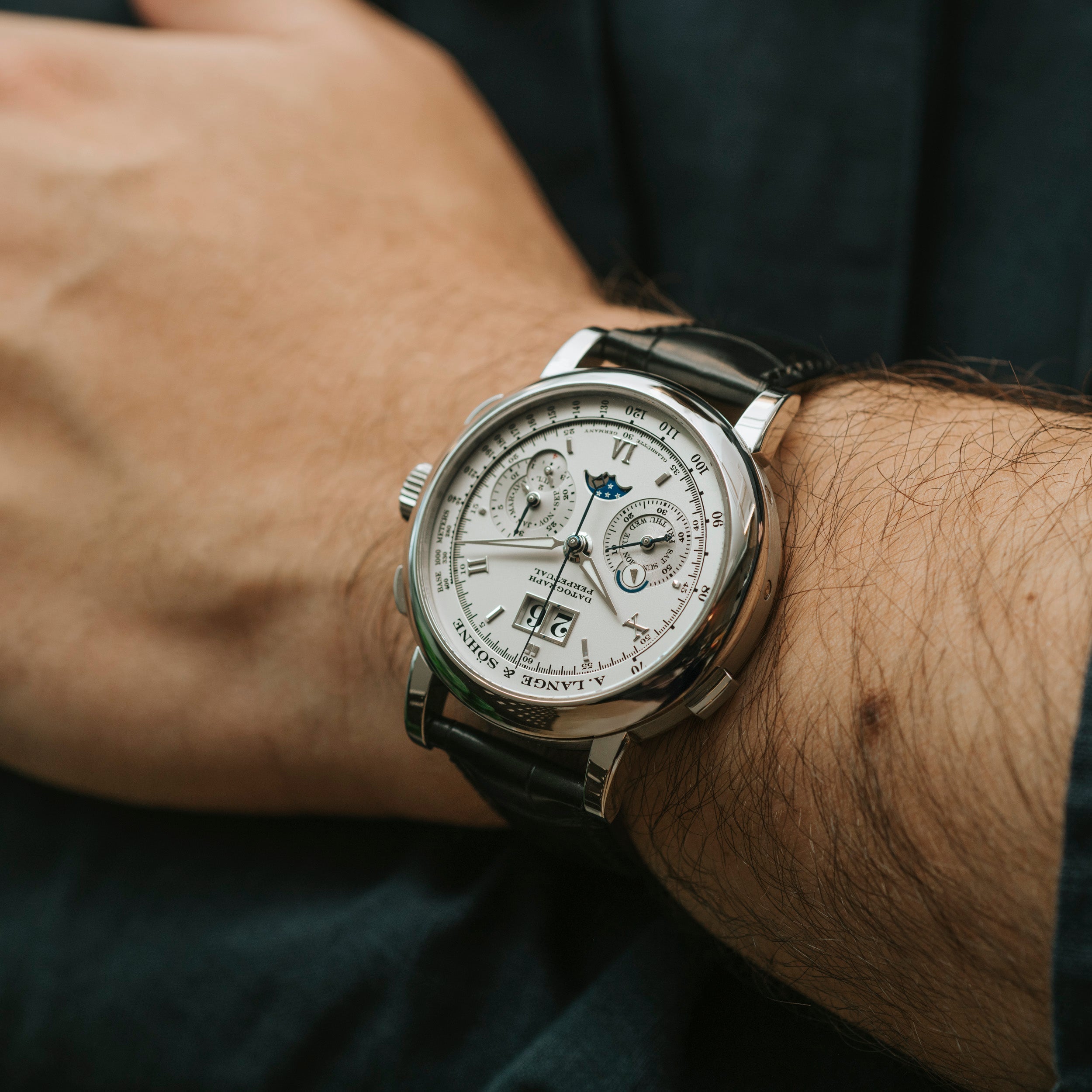 The Lange & Sohne Datograph Perpetual Calendar, a luxury wristwatch with a white dial and black leather strap, is shown on a person’s wrist.