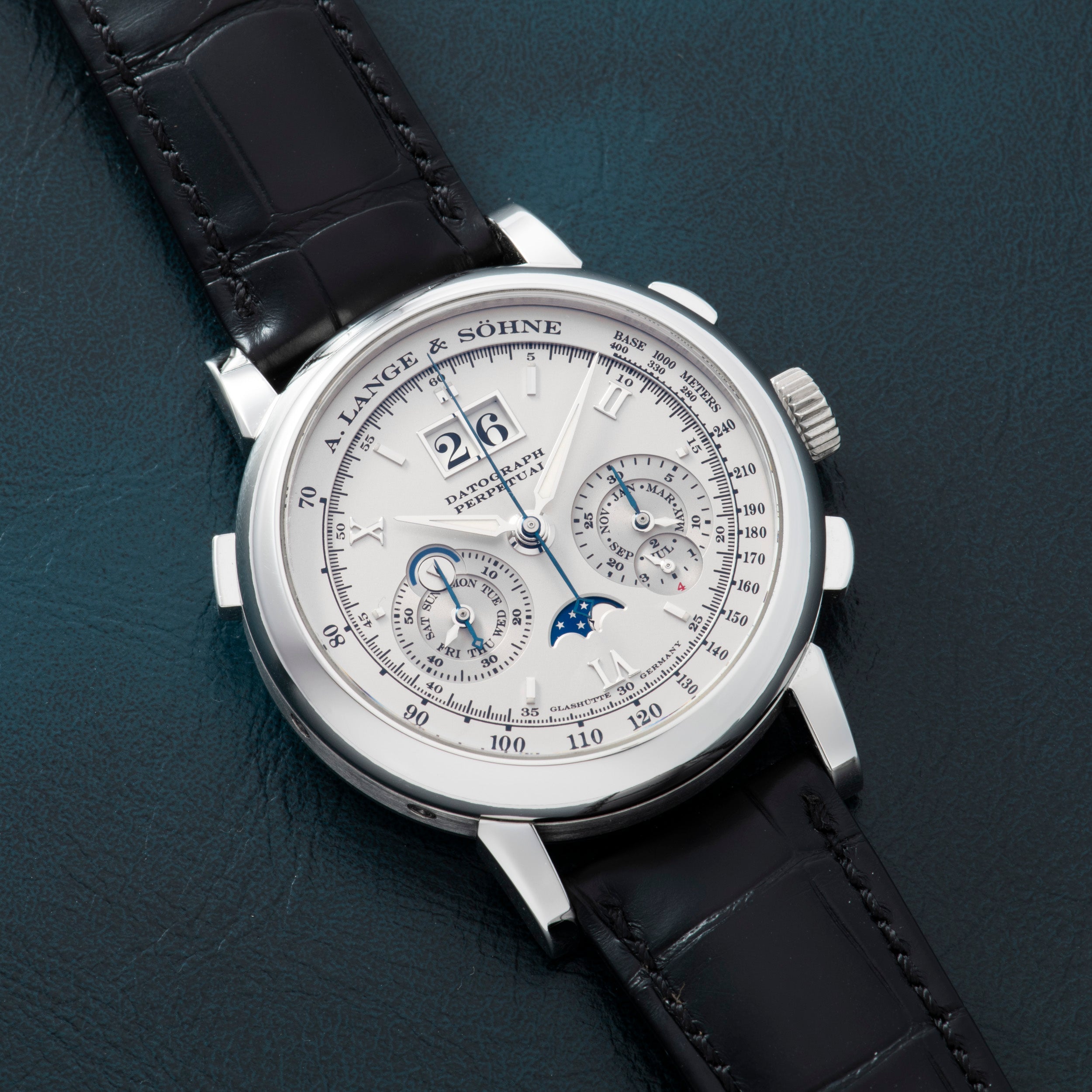 A Lange & Sohne Datograph Perpetual Calendar wristwatch with a silver dial and black leather strap displayed on a dark teal surface.