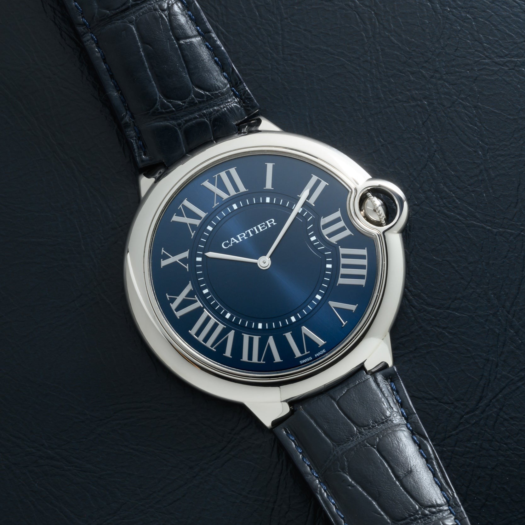 A Cartier Ballon Bleu watch with a blue dial and Roman numerals on a black leather strap, displayed on a dark textured surface.