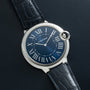 A Cartier Ballon Bleu watch with a blue dial and Roman numerals on a black leather strap, displayed on a dark textured surface.