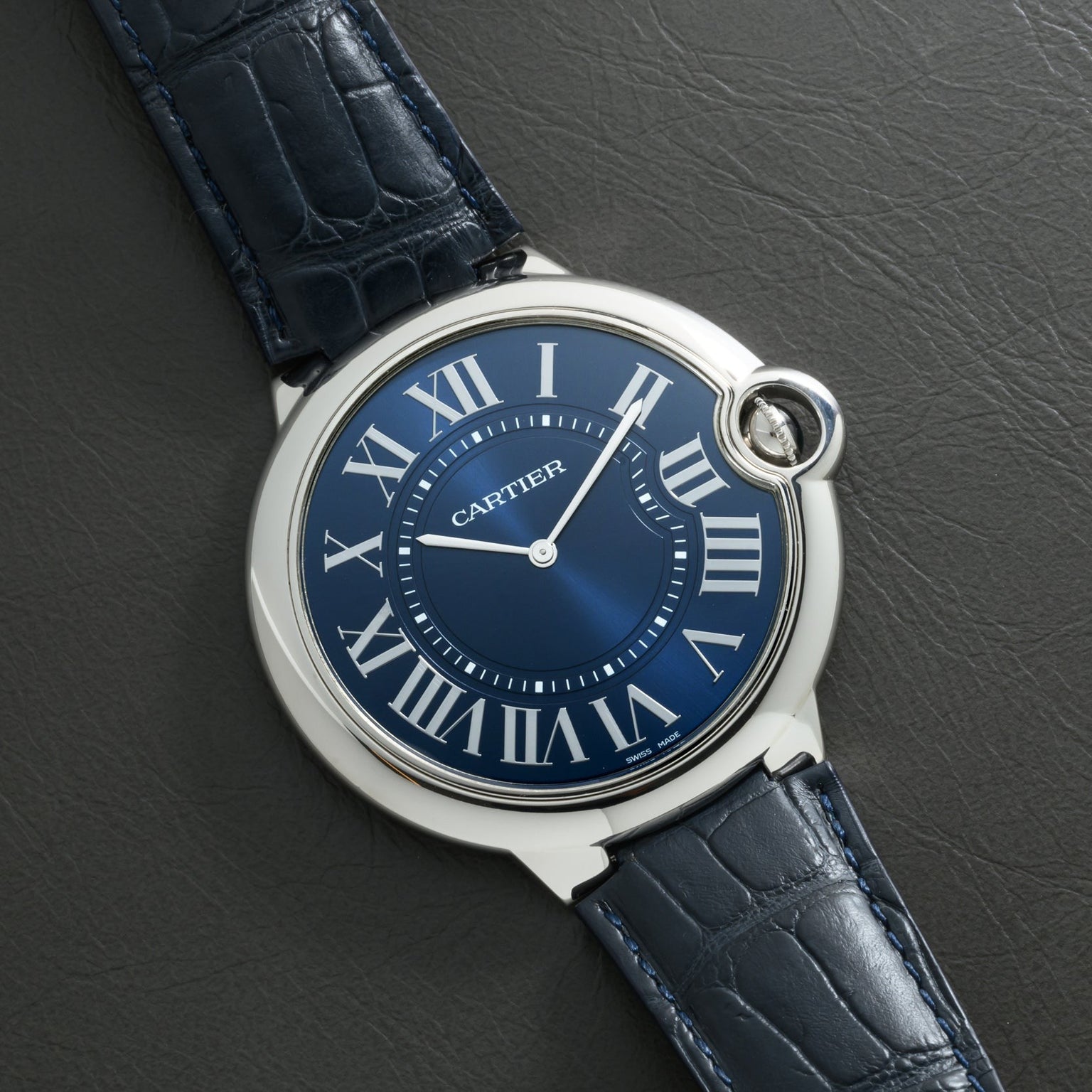 A Cartier Ballon Bleu watch with Roman numerals and a leather strap rests on a textured dark surface.
