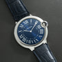 A Cartier Ballon Bleu watch with Roman numerals and a leather strap rests on a textured dark surface.