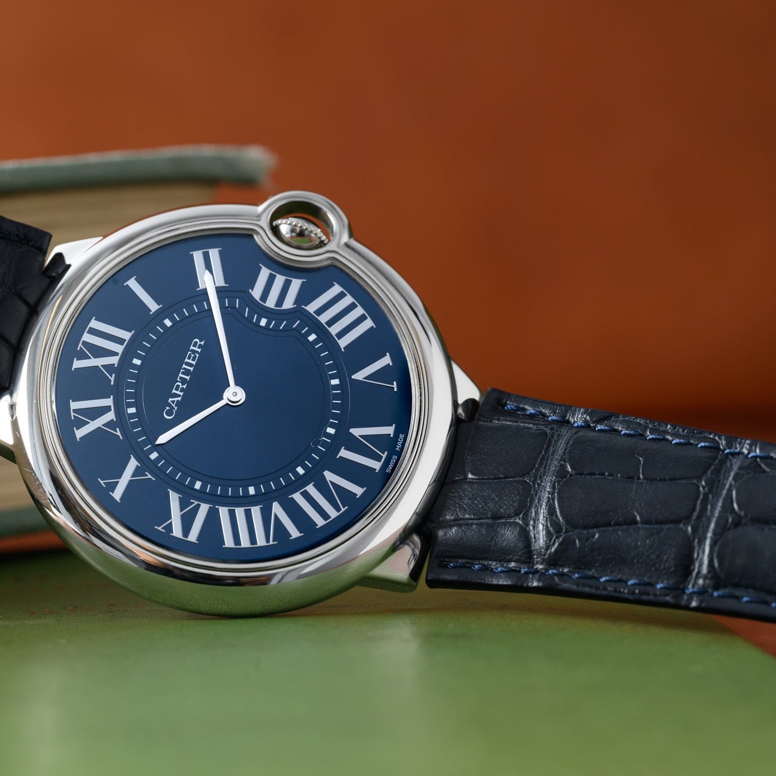 A Cartier Ballon Bleu watch with a blue face, Roman numerals, and a black leather strap rests on a green surface.