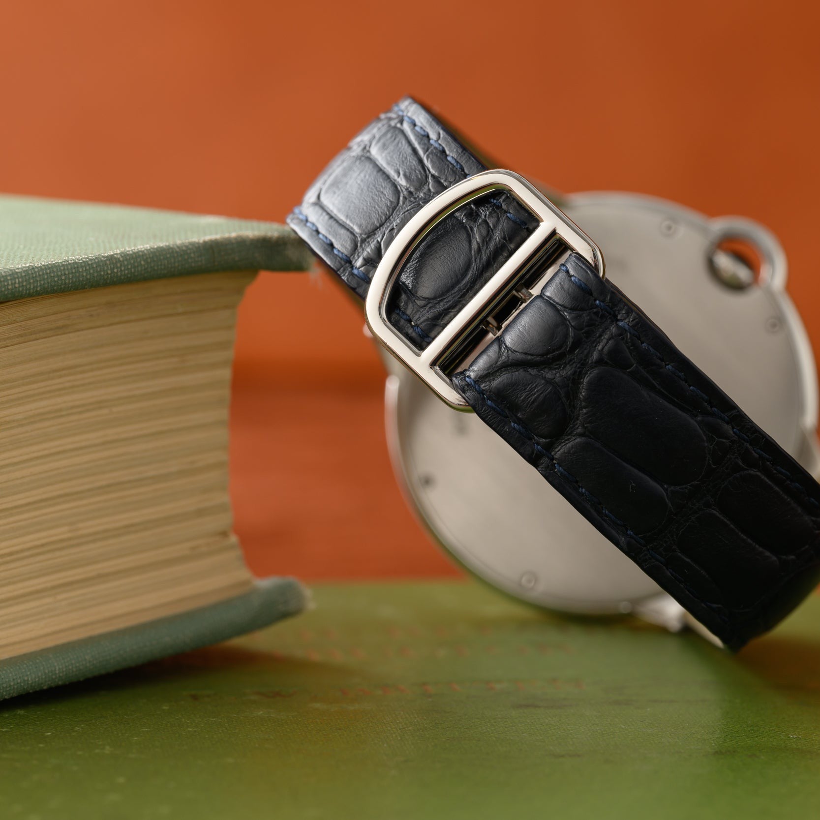 A Cartier Ballon Bleu black leather watch strap rests on a closed book, set against a blurred orange background.