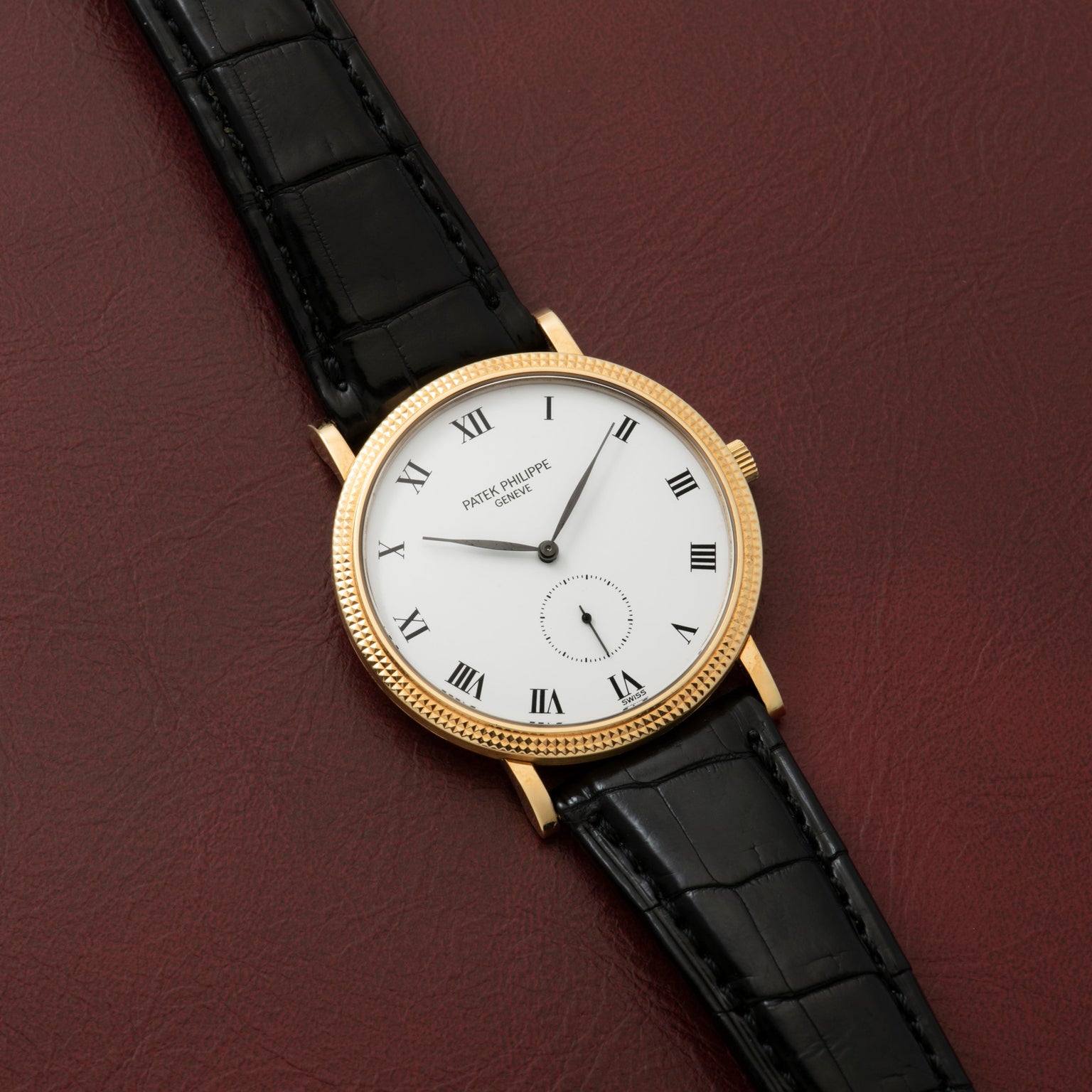 Patek Philippe Calatrava gold watch with black leather strap and Roman numerals displayed on a burgundy leather surface.
