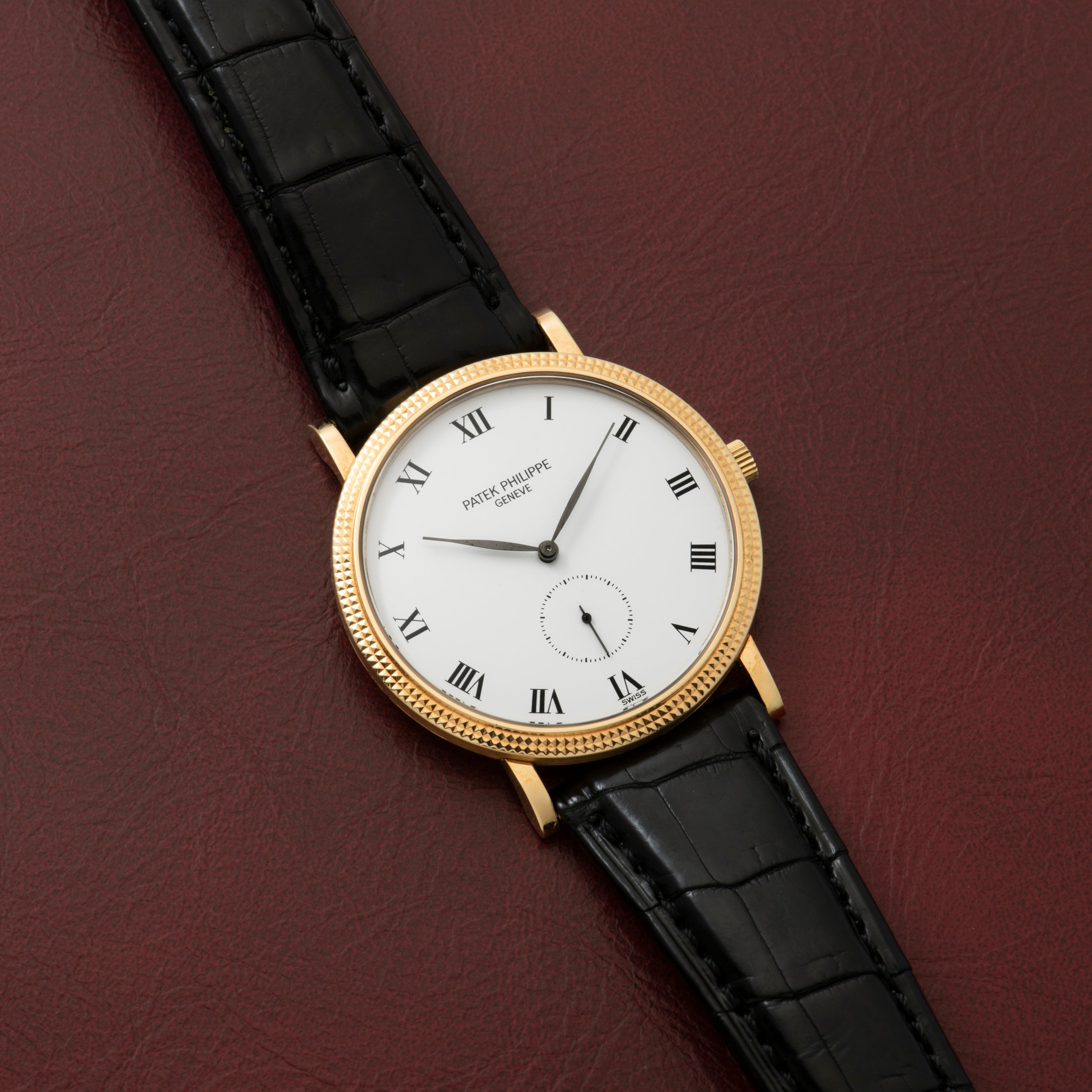 Patek Philippe Calatrava gold watch with black leather strap and Roman numerals displayed on a burgundy leather surface.