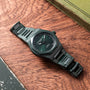 The Laureato Green Ceramic Aston Martin Edition by Girard-Perregaux, with its textured green dial, rests on a wooden surface near a green book.
