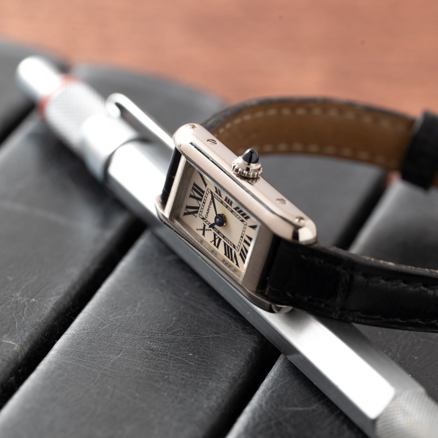 The Cartier Tank Ladies Allongee wristwatch with a black leather strap rests on a silver pen atop a black notebook.