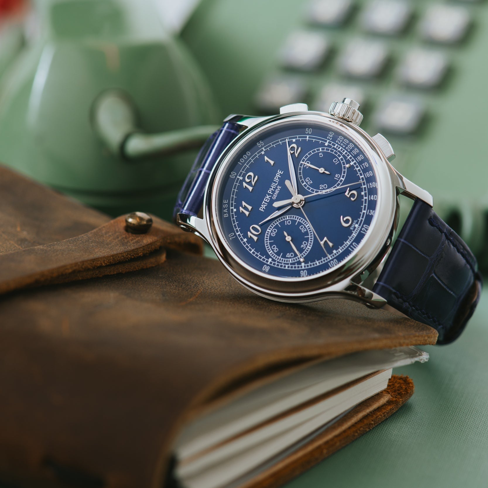 The Patek Philippe Split-Seconds Chronograph with a blue face sits atop a leather notebook beside a vintage green rotary phone.