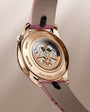 Rose gold watch with visible skeleton back displaying mechanical movement, purple and taupe leather strap with black accents.