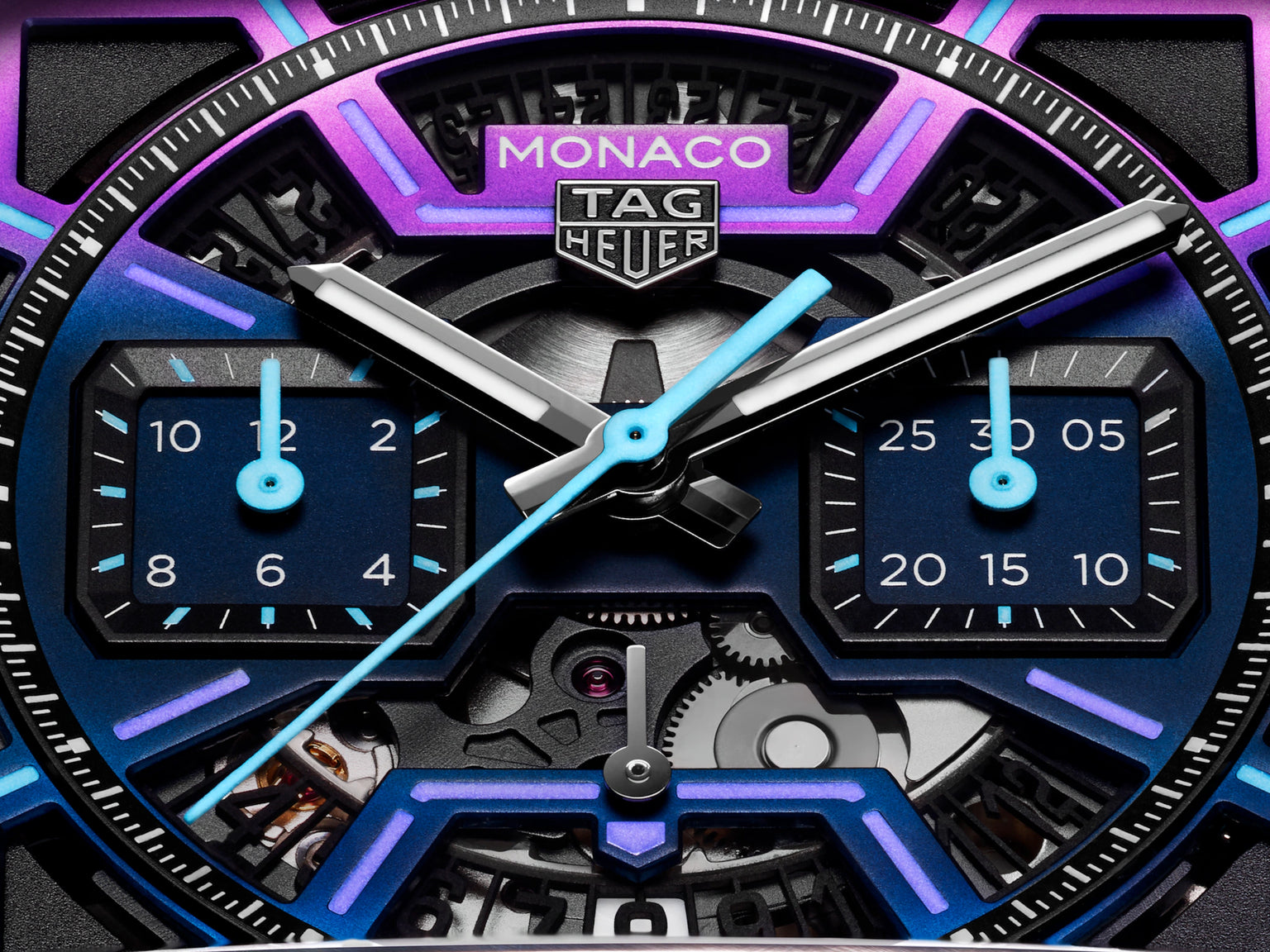 Close-up of the TAG Heuer Monaco Chronograph Las Vegas Edition watch face, featuring a geometric blue and purple design, square subdials, exposed gears, and a sleek titanium DLC-coated case. TAG Heuer logo appears at the top center.