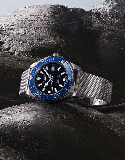 Watch with blue bezel and silver mesh band on a rocky surface