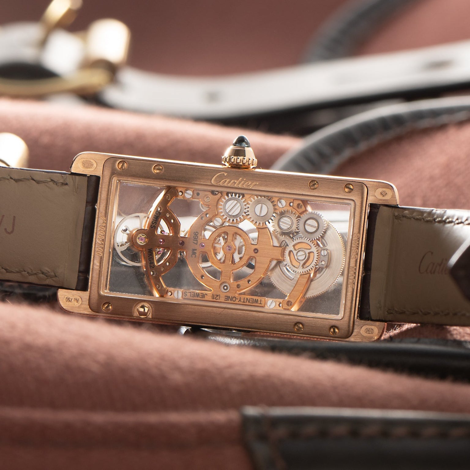 A close-up of the Cartier Tank Cintreé Skeleton watch, featuring exposed gears and a leather strap, resting on brown fabric.
