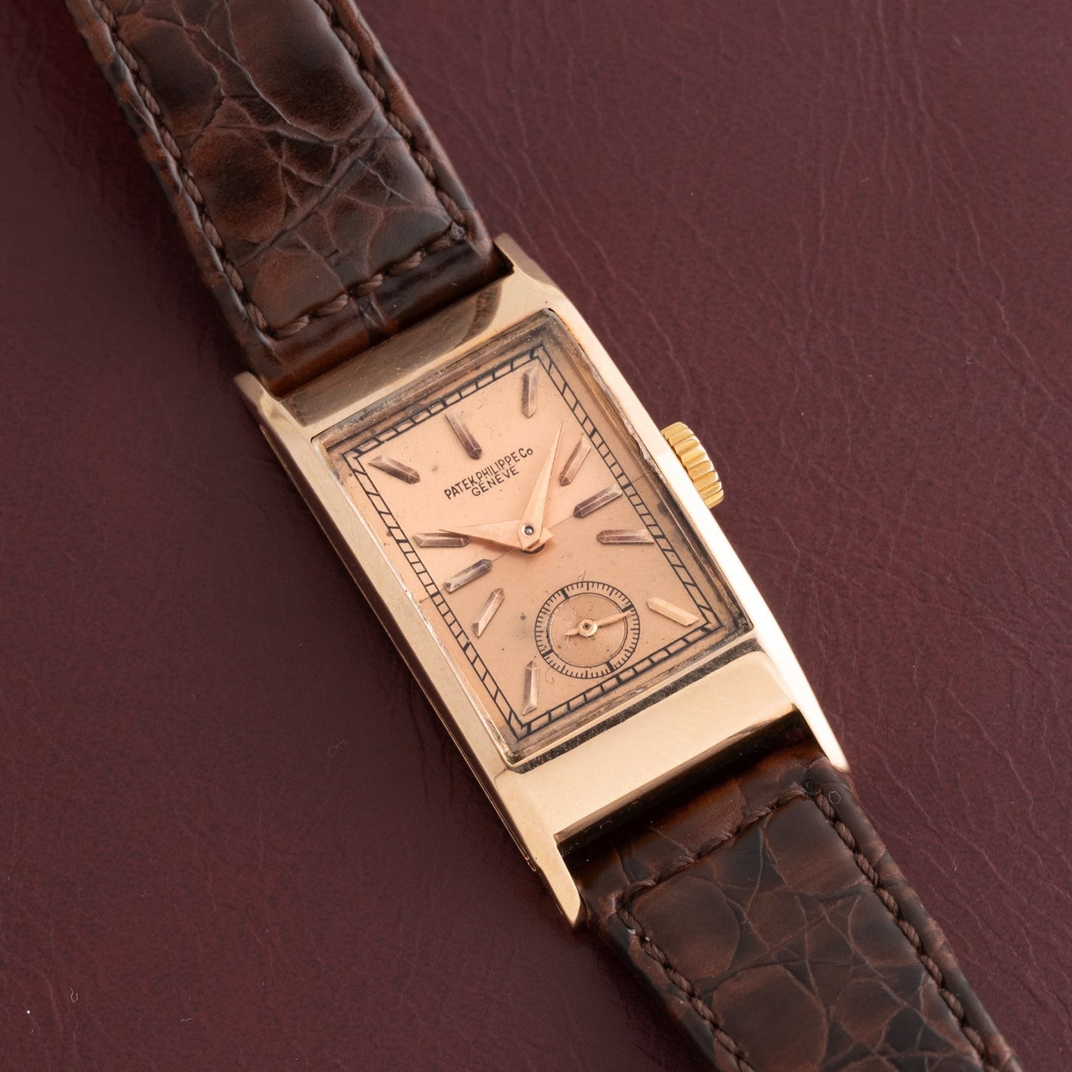 The Patek Philippe Tegolino Dress Watch, featuring a rectangular gold case and brown leather strap, displayed on a dark burgundy leather surface.
