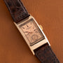 The Patek Philippe Tegolino Dress Watch, featuring a rectangular gold case and brown leather strap, rests on a brown leather surface.