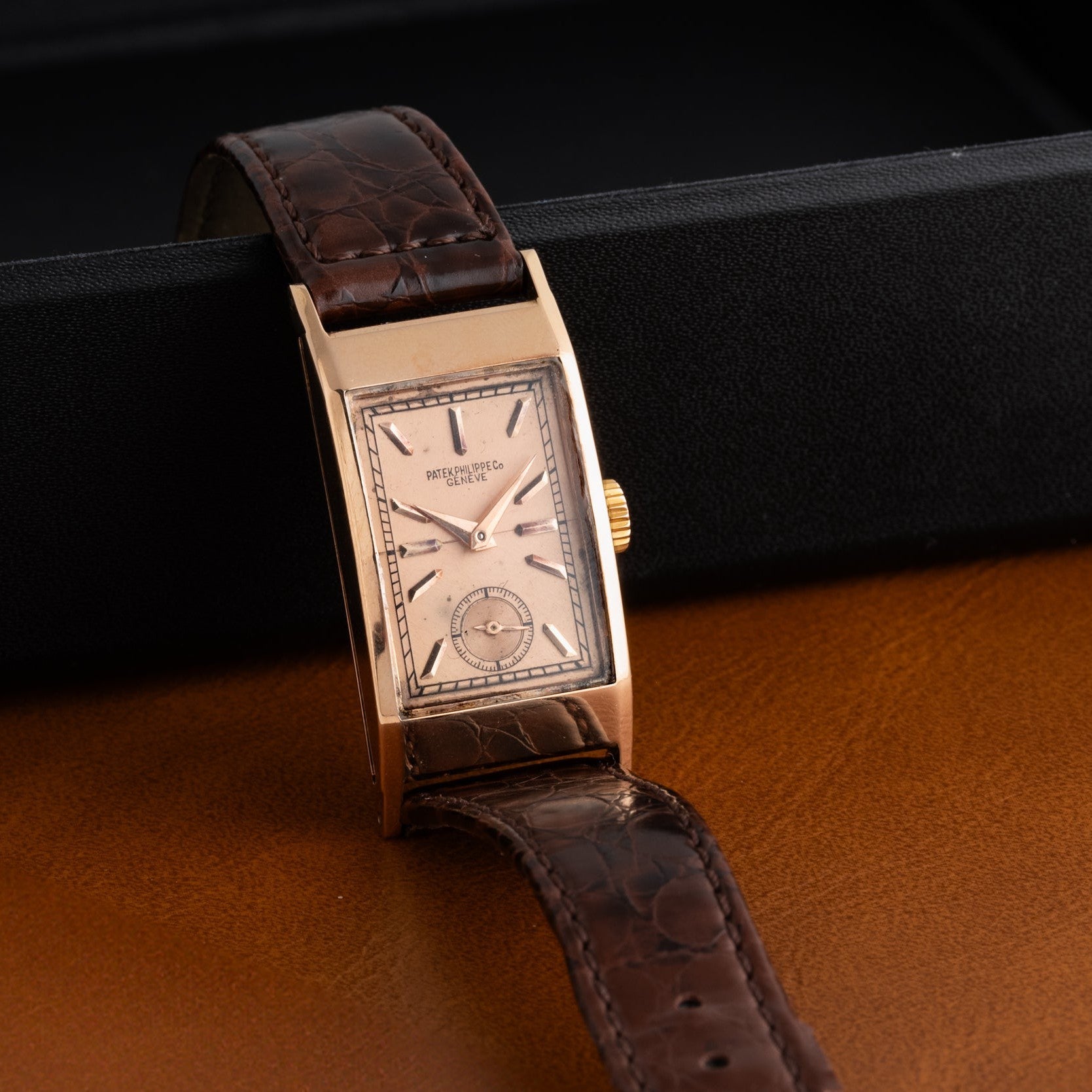 The Patek Philippe Tegolino Dress Watch features a vintage rectangular gold case and a brown leather strap, elegantly displayed on a black and brown surface.
