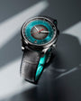 Stainless steel watch with teal dial, chronograph subdial, red hands, black leather strap, and teal inner band.