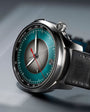 Silver chronograph watch with teal dial, center subdial, red chronograph hand, and black leather strap.