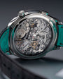 Silver chronograph watch with visible skeleton movement, teal leather strap, and textured crown against gray background.
