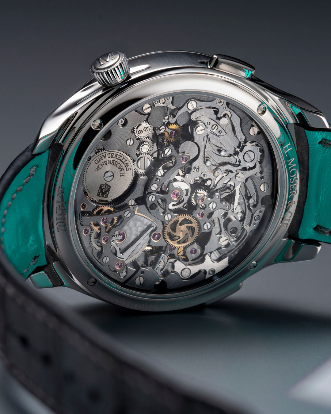 Silver chronograph watch with visible skeleton movement, teal leather strap, and textured crown against gray background.